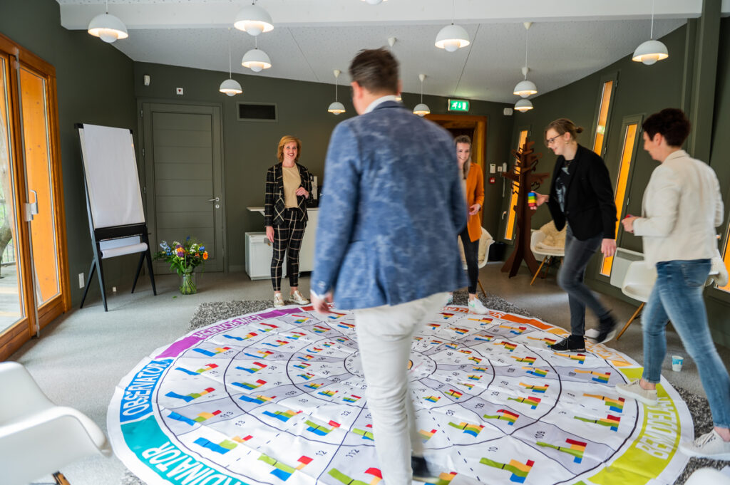 Een teamcoach staat bij een Insights Discovery-kleurenwiel en legt iets uit aan een groep mensen die aandachtig luisteren. De sfeer is informeel en betrokken, passend bij een interactieve workshop over persoonlijkheidstypes en teamdynamiek